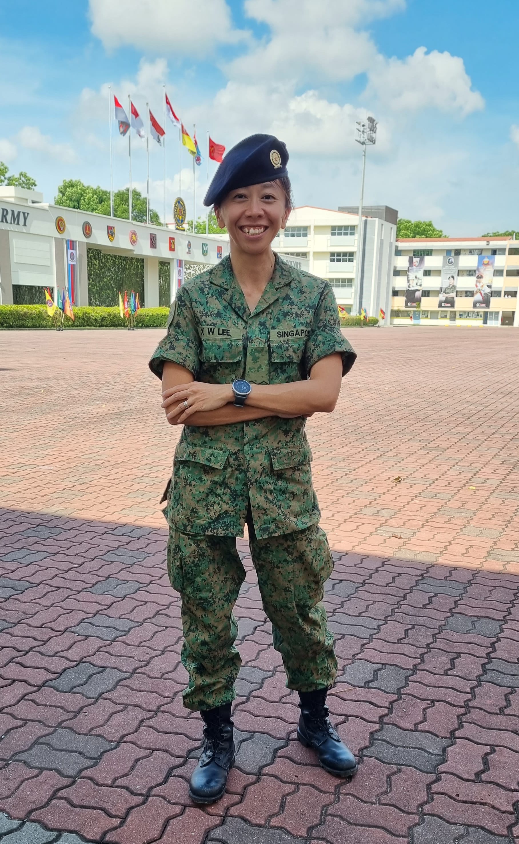 Person in camouflage uniform stands with arms crossed wearing beret and watch, outdoor flags in background.